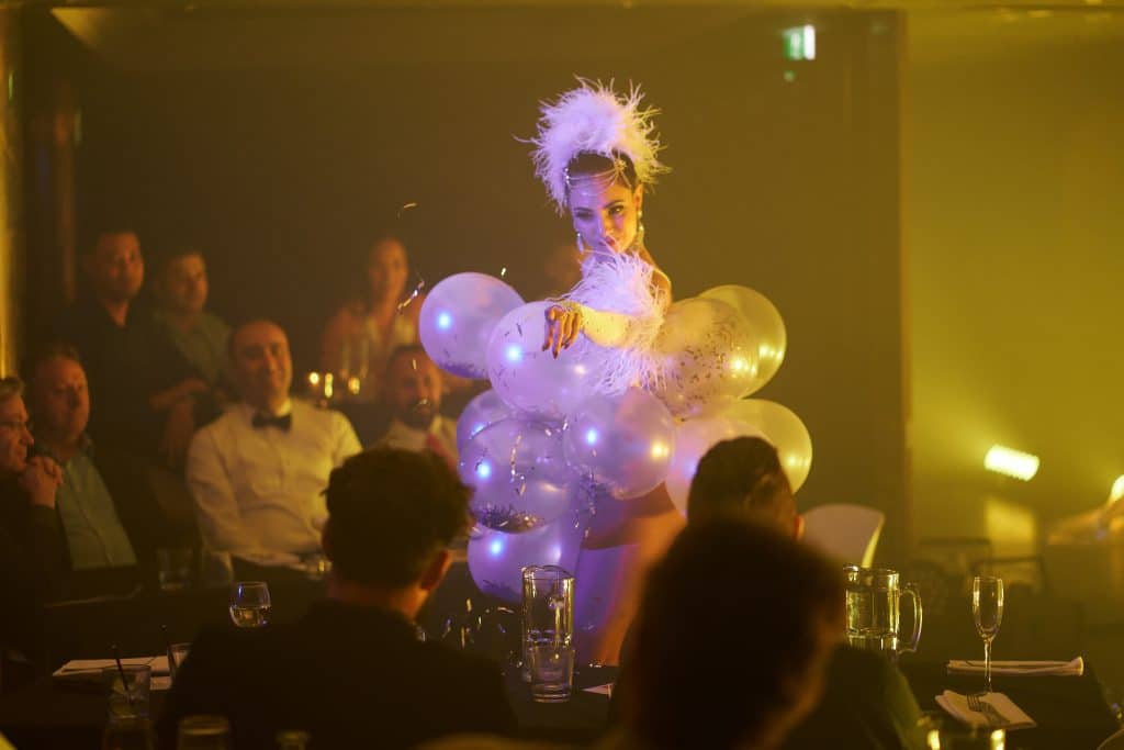 female variety show performer dressed up in balloon outfit for talk and tease performance, on stage pointing towards a member of the audience