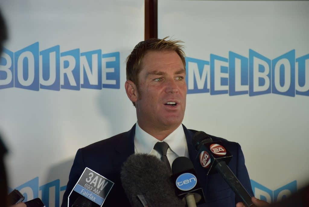 shane warne in front of cameras and a sign of melbourne
