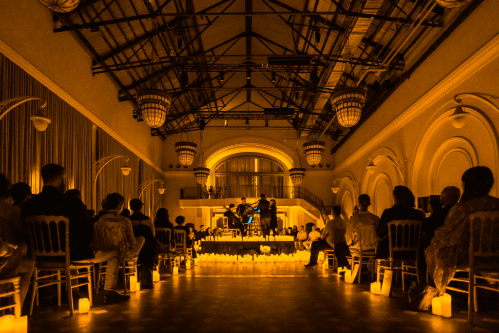 A shot taken from the audience capturing the beams and low hanging lights above a stage covered in the glow of candlelight at a Candlelight concert inside The Eveleigh in Sydney.