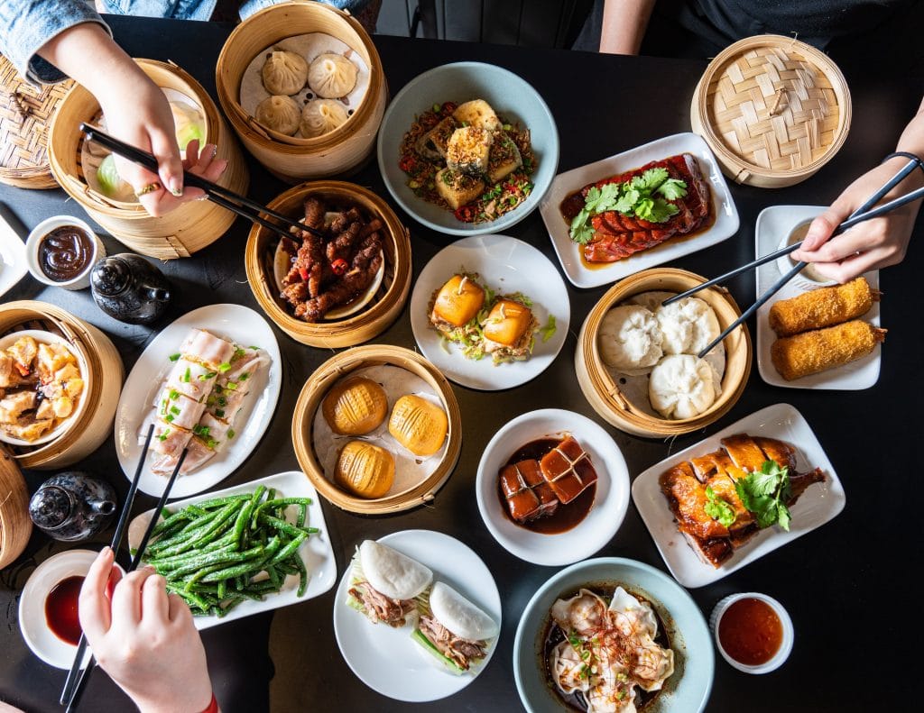 bird's eye view of a selection of yum cha dishes like dumplings and more with people picking at the food with chop sticks