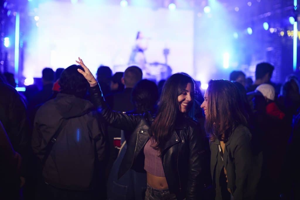 crowded concert room, music on stage behind crowd, two young women at forefront