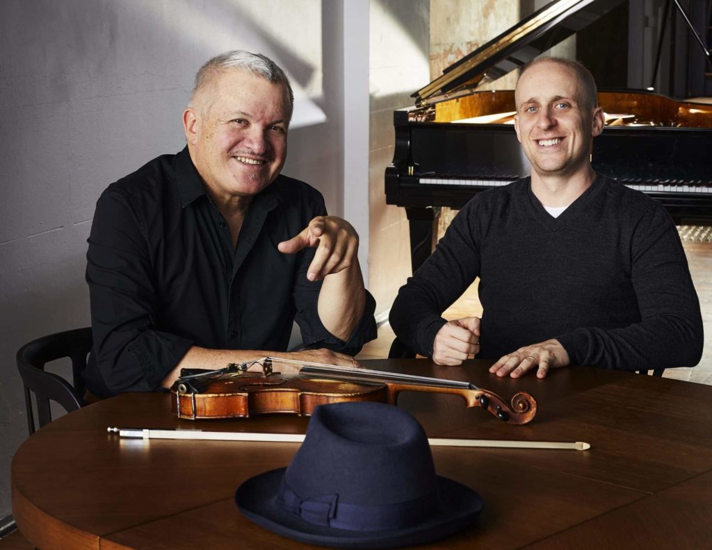 george washingmachine and simon tedeschi seated at a table with a violin laying on it and a grand piano in the background