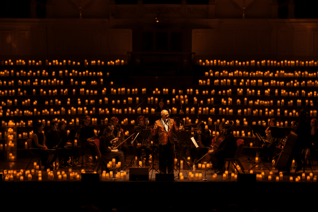 A conductor standing in the centre of a stage in front of an orchestra and wall of candles.