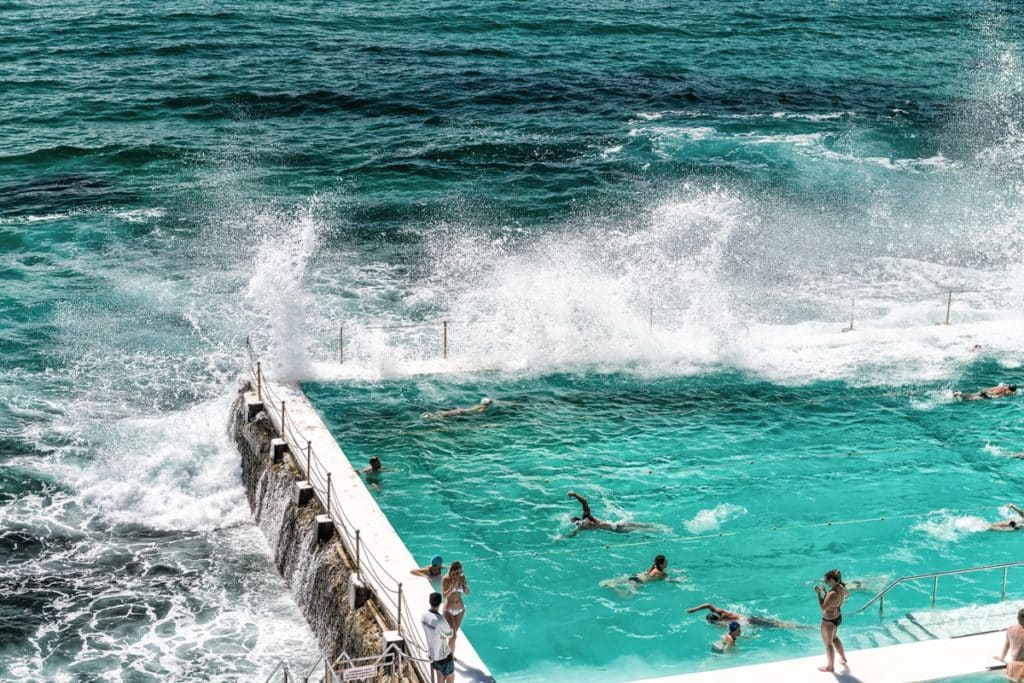 Ocean waves crash against the edge of Bondi Icebergs pool, filled with swimmers.