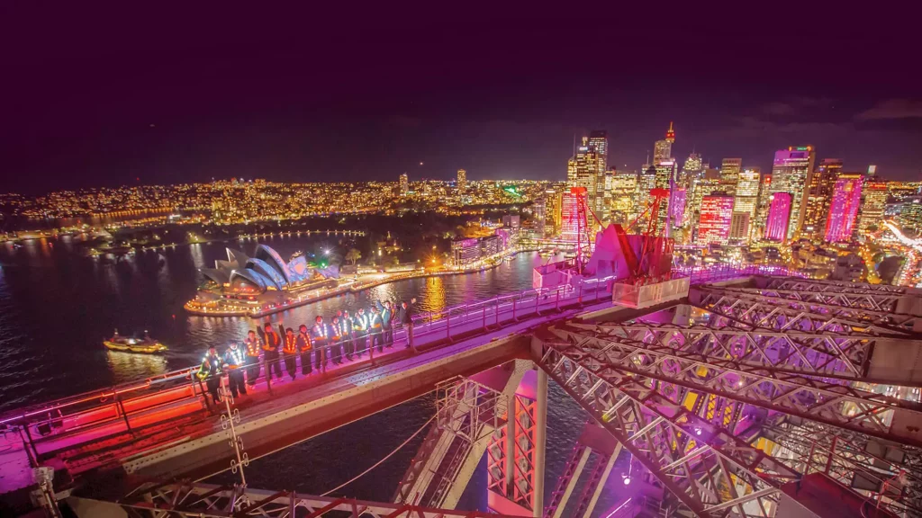 people climbing the harbour bridge with vivid sydney lights around the harbour
